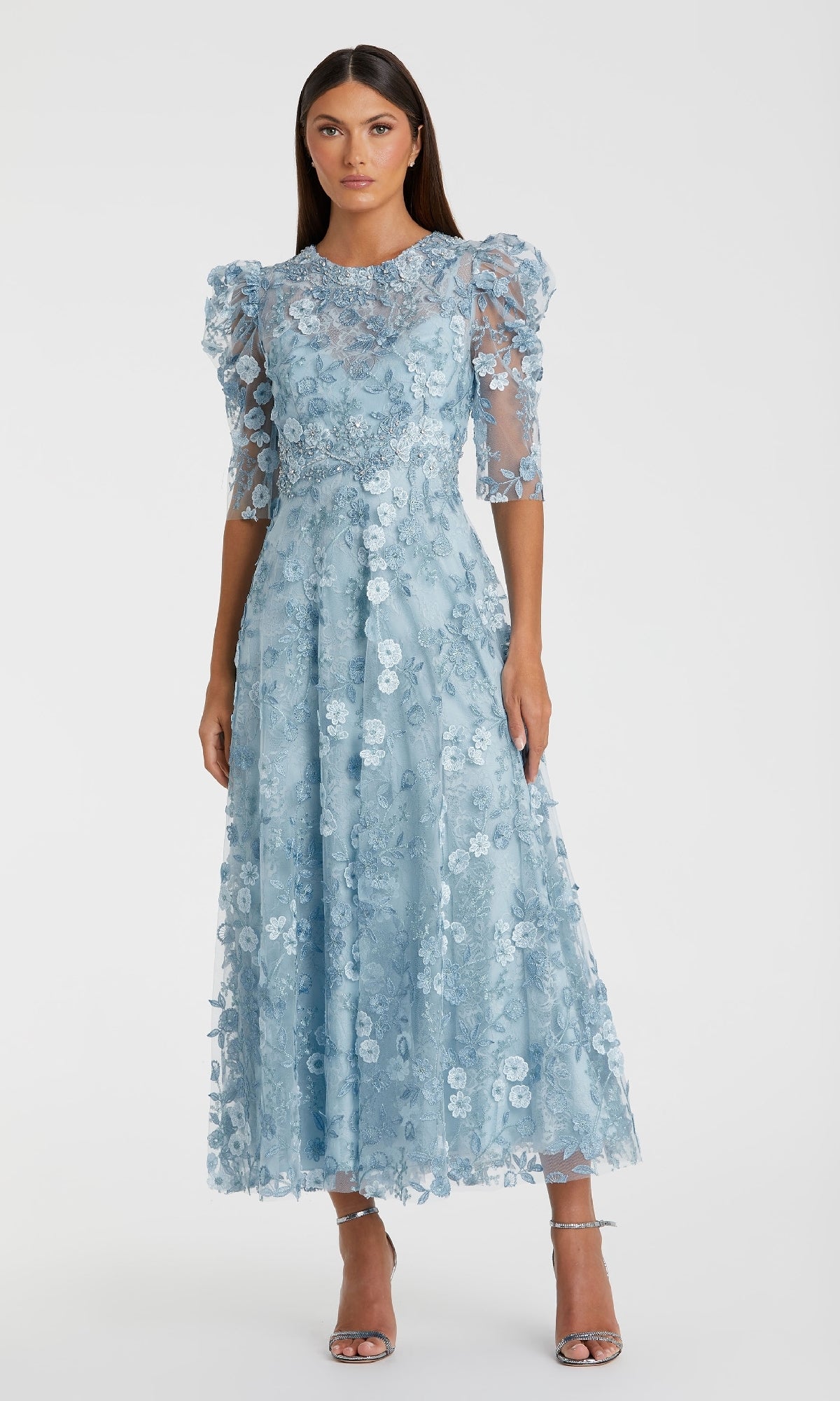 A woman wears the Mac Duggal 68193 tea-length lace formal dress in light blue, featuring puffed sleeves and sheer details, styled with silver high-heeled sandals.